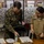 Two military personnel, one in US Marine Corps digital camouflage uniform and one in a tan/khaki uniform, are standing at a desk reviewing documents. The Marine, identified by name tape as 'KLOTZ,' appears to be presenting or signing certificates alongside the other service member identified as 'FAUNI.' The setting appears to be a military office or administrative area.