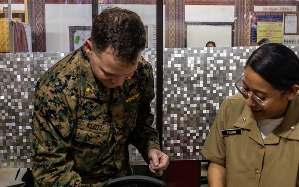 Two military personnel, one in US Marine Corps digital camouflage uniform and one in a tan/khaki uniform, are standing at a desk reviewing documents. The Marine, identified by name tape as 'KLOTZ,' appears to be presenting or signing certificates alongside the other service member identified as 'FAUNI.' The setting appears to be a military office or administrative area.
