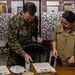 Two military personnel, one in US Marine Corps digital camouflage uniform and one in a tan/khaki uniform, are standing at a desk reviewing documents. The Marine, identified by name tape as 'KLOTZ,' appears to be presenting or signing certificates alongside the other service member identified as 'FAUNI.' The setting appears to be a military office or administrative area.