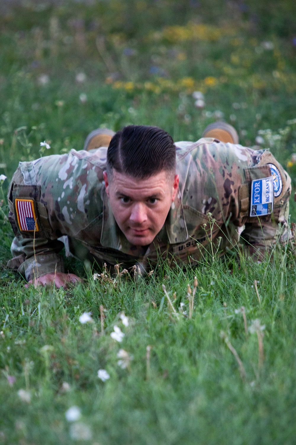 A U.S. Army soldier in OCP (Operational Camouflage Pattern) uniform is performing a low crawl exercise on a grassy field with wildflowers. The soldier is looking directly at the camera with a determined expression. Visible patches indicate affiliation with KFOR RC-East, suggesting a deployment or training exercise in Kosovo.