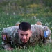 A U.S. Army soldier in OCP (Operational Camouflage Pattern) uniform is performing a low crawl exercise on a grassy field with wildflowers. The soldier is looking directly at the camera with a determined expression. Visible patches indicate affiliation with KFOR RC-East, suggesting a deployment or training exercise in Kosovo.