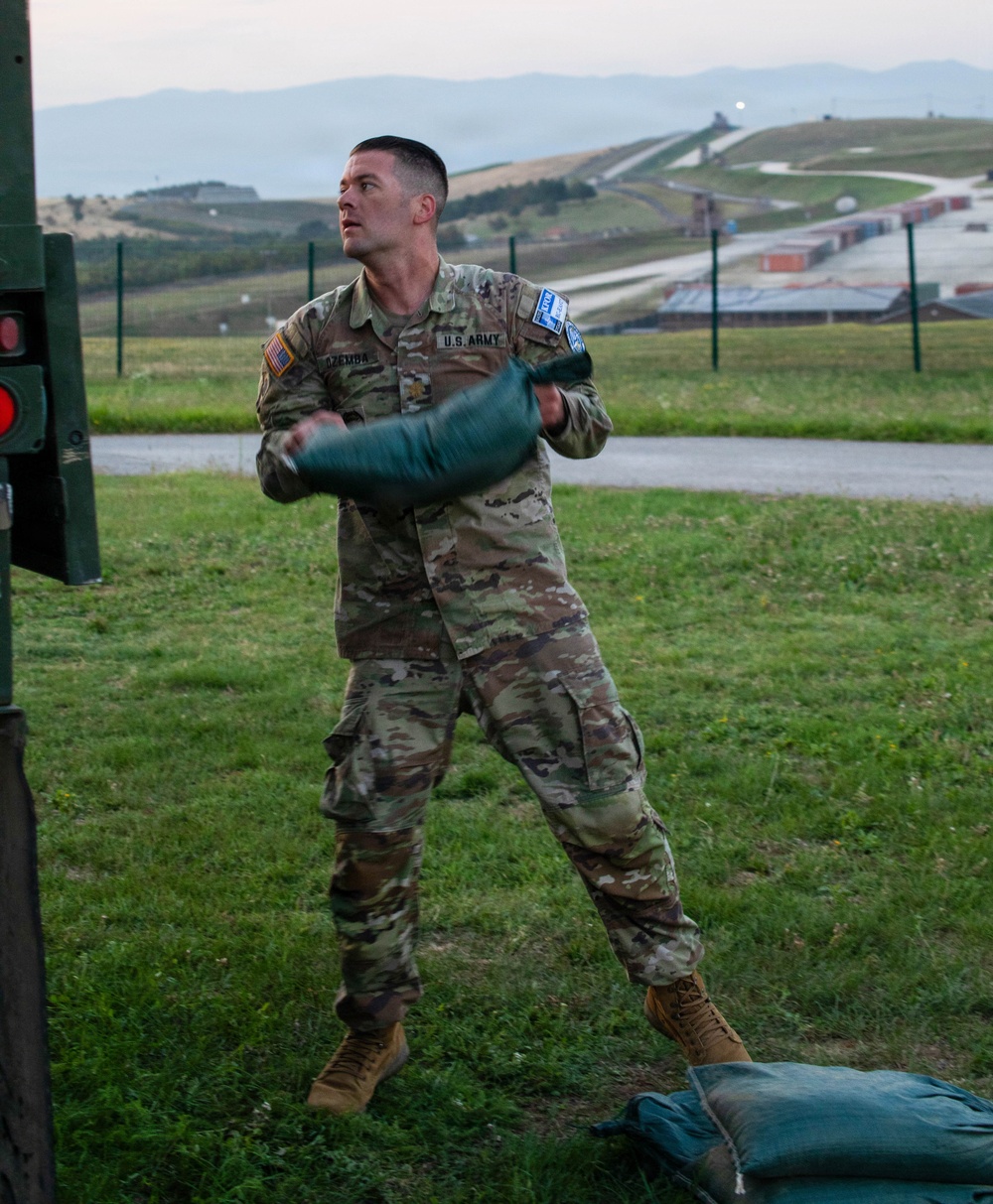 A U.S. Army soldier in OCP (Operational Camouflage Pattern) uniform is actively throwing or tossing a green sandbag, likely during a training exercise or competition. The soldier's name tape reads 'DZEMBA' and he wears a U.S. flag patch on his left shoulder along with what appears to be a competition bib. The setting appears to be a military installation with rolling hills, a road, and shipping containers visible in the background.