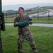 A U.S. Army soldier in OCP (Operational Camouflage Pattern) uniform is actively throwing or tossing a green sandbag, likely during a training exercise or competition. The soldier's name tape reads 'DZEMBA' and he wears a U.S. flag patch on his left shoulder along with what appears to be a competition bib. The setting appears to be a military installation with rolling hills, a road, and shipping containers visible in the background.