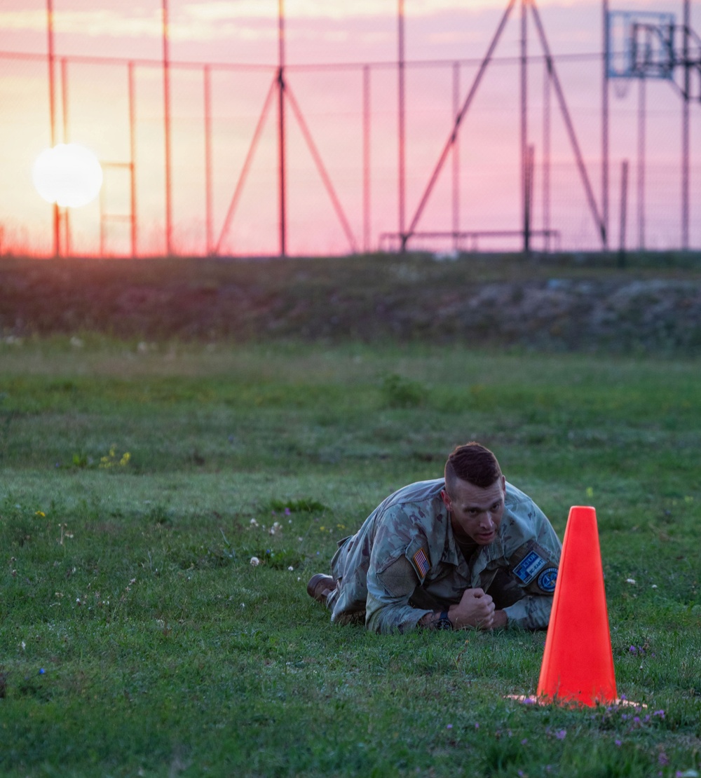 A soldier in U.S. Army camouflage uniform is performing a low crawl exercise on a grass field at either sunrise or sunset. The soldier is prone on the ground, moving past an orange traffic cone, likely as part of a physical fitness or obstacle course training event. The dramatic sky with warm orange and pink hues provides a striking backdrop against what appears to be a military training facility.