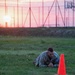 A soldier in U.S. Army camouflage uniform is performing a low crawl exercise on a grass field at either sunrise or sunset. The soldier is prone on the ground, moving past an orange traffic cone, likely as part of a physical fitness or obstacle course training event. The dramatic sky with warm orange and pink hues provides a striking backdrop against what appears to be a military training facility.