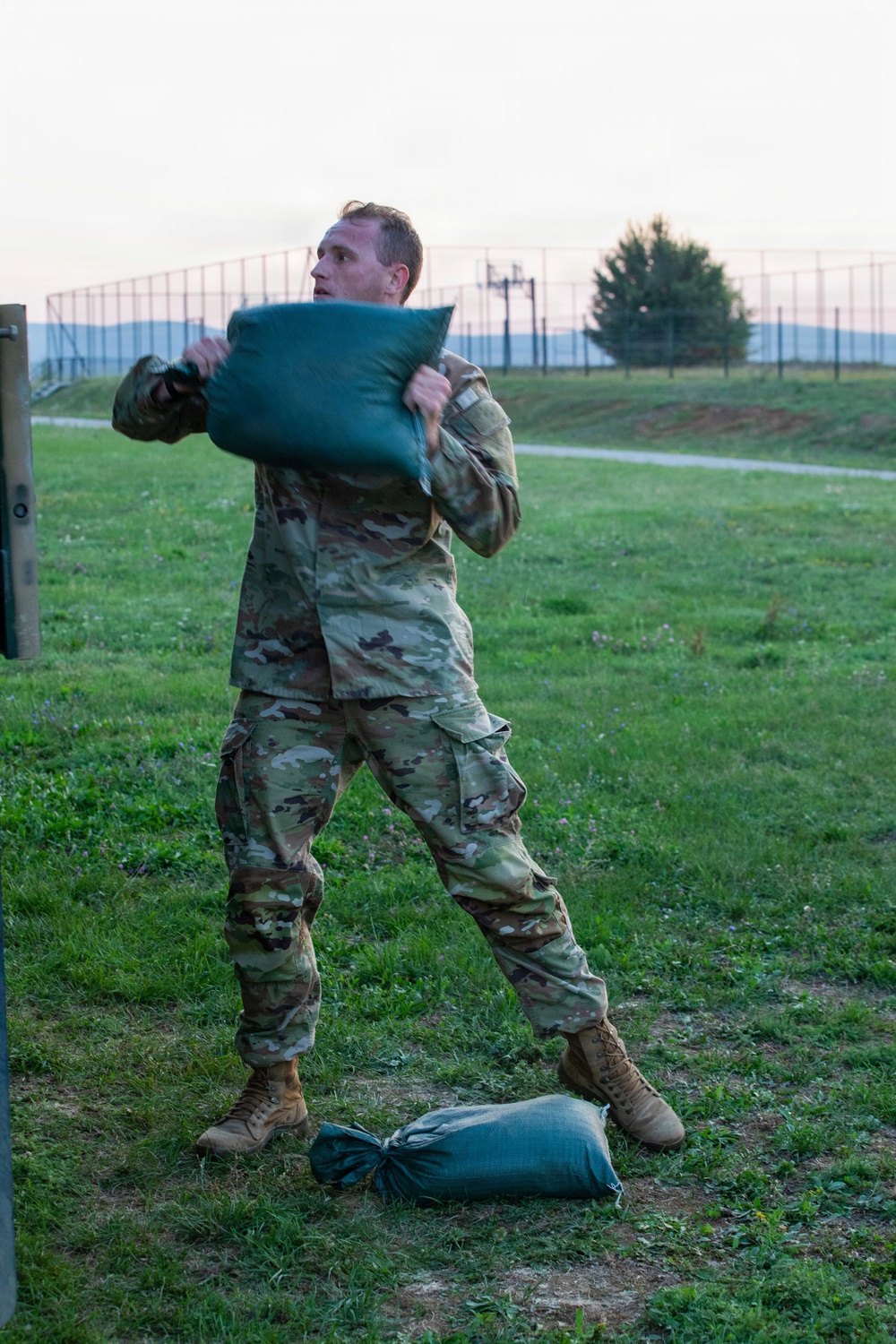 A soldier in US Army OCP (Operational Camouflage Pattern) uniform is lifting or throwing a large green sandbag during what appears to be a physical training or obstacle course exercise. The activity takes place on a grassy field at dusk or dawn, with a fenced perimeter and trees visible in the background. A second sandbag lies on the ground near his feet.