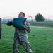 A soldier in US Army OCP (Operational Camouflage Pattern) uniform is lifting or throwing a large green sandbag during what appears to be a physical training or obstacle course exercise. The activity takes place on a grassy field at dusk or dawn, with a fenced perimeter and trees visible in the background. A second sandbag lies on the ground near his feet.