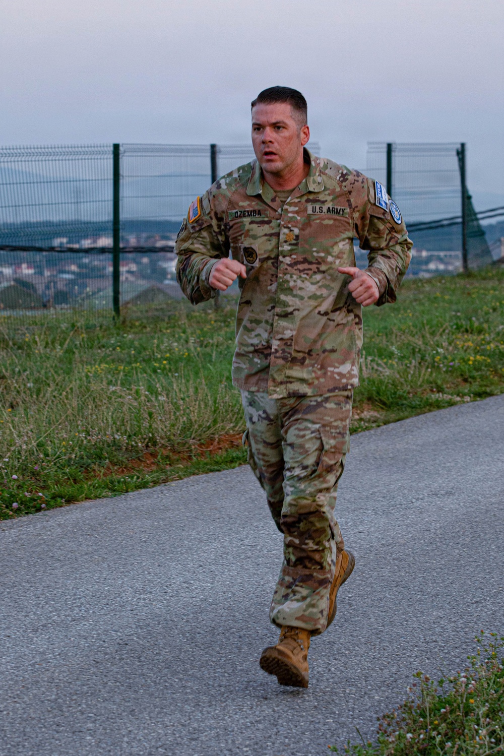 A U.S. Army soldier named DZEMBA, wearing OCP (Operational Camouflage Pattern) uniform, is running along a paved path near a perimeter fence. He appears to be conducting a physical fitness run, likely as part of a physical training exercise or fitness test. The setting appears to be an overseas military installation based on the visible patches and surrounding landscape.
