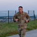 A U.S. Army soldier named DZEMBA, wearing OCP (Operational Camouflage Pattern) uniform, is running along a paved path near a perimeter fence. He appears to be conducting a physical fitness run, likely as part of a physical training exercise or fitness test. The setting appears to be an overseas military installation based on the visible patches and surrounding landscape.