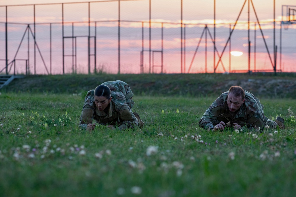 Two military personnel in camouflage uniforms are performing a low crawl exercise on a grass field during sunset or sunrise. They are both prone on the ground, moving forward on their elbows and knees in what appears to be a physical training or tactical drill. A chain-link fence and athletic facility structures are visible in the background against a pink and orange sky.