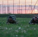 Two military personnel in camouflage uniforms are performing a low crawl exercise on a grass field during sunset or sunrise. They are both prone on the ground, moving forward on their elbows and knees in what appears to be a physical training or tactical drill. A chain-link fence and athletic facility structures are visible in the background against a pink and orange sky.
