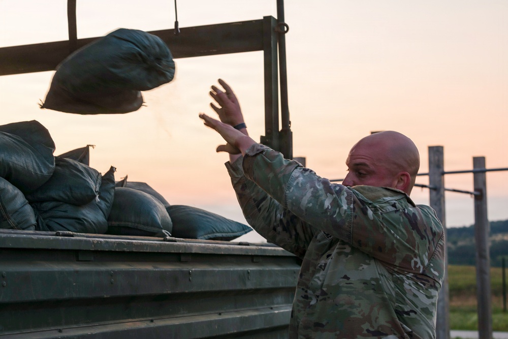 A bald soldier in camouflage uniform is tossing sandbags onto a military truck bed at dusk or dawn. The scene appears to be a field operation involving the loading or unloading of sandbags, likely for flood control, fortification, or base preparation. The warm orange sky in the background suggests early morning or evening lighting conditions.
