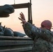 A bald soldier in camouflage uniform is tossing sandbags onto a military truck bed at dusk or dawn. The scene appears to be a field operation involving the loading or unloading of sandbags, likely for flood control, fortification, or base preparation. The warm orange sky in the background suggests early morning or evening lighting conditions.