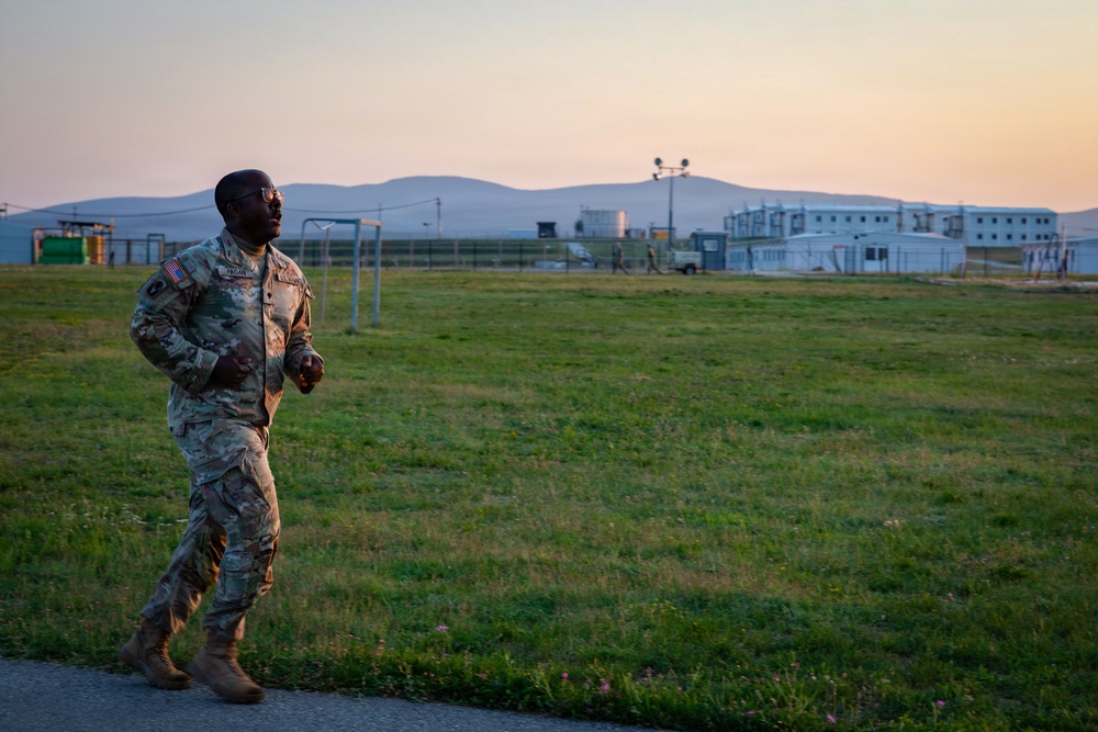 A U.S. Army soldier in MultiCam/OCP camouflage uniform is jogging along a path at what appears to be a military installation during early morning or evening hours. The soldier wears glasses and has a U.S. flag patch visible on the left shoulder. The background shows modular military buildings, a fenced compound, and rolling hills under a pastel-colored sky.