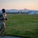 A U.S. Army soldier in MultiCam/OCP camouflage uniform is jogging along a path at what appears to be a military installation during early morning or evening hours. The soldier wears glasses and has a U.S. flag patch visible on the left shoulder. The background shows modular military buildings, a fenced compound, and rolling hills under a pastel-colored sky.