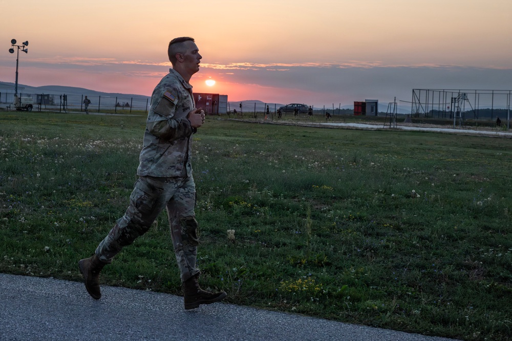 A U.S. Army soldier in full camouflage uniform and combat boots is running along a paved path at what appears to be a military installation during sunset. The warm orange and pink sky frames the scene as the soldier runs in profile view. In the background, other personnel, vehicles, shipping containers, and sports field structures are visible across an open grassy area.