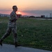 A U.S. Army soldier in full camouflage uniform and combat boots is running along a paved path at what appears to be a military installation during sunset. The warm orange and pink sky frames the scene as the soldier runs in profile view. In the background, other personnel, vehicles, shipping containers, and sports field structures are visible across an open grassy area.