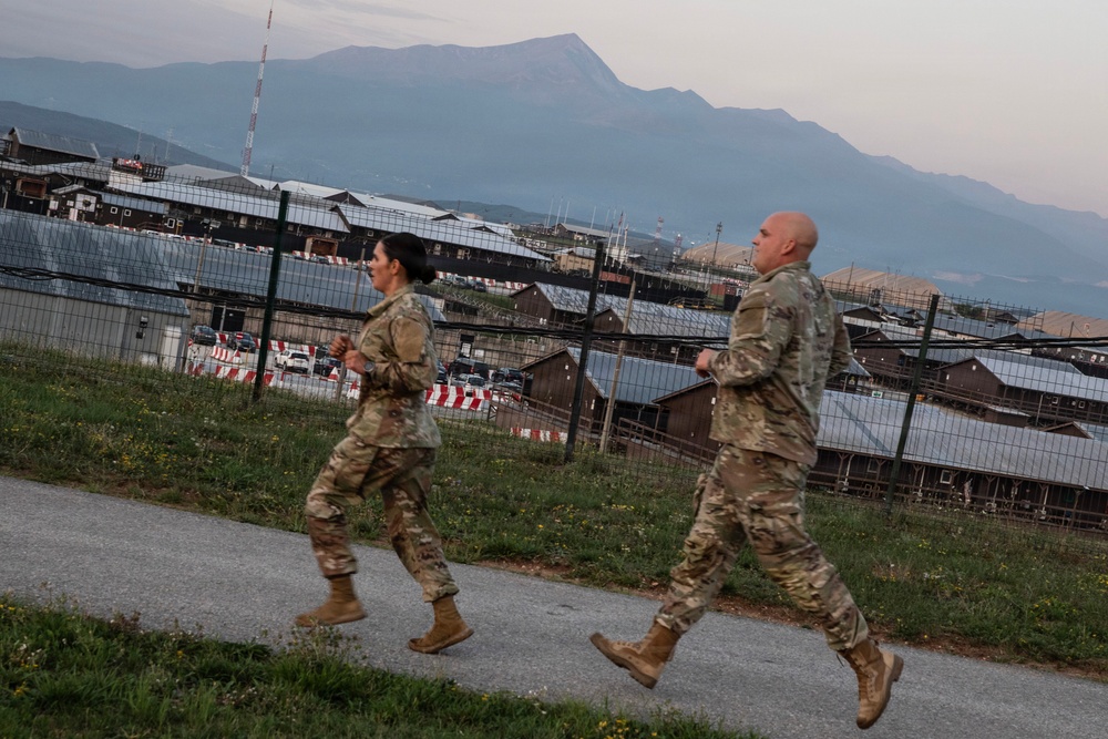 Two military personnel in camouflage uniforms and tan combat boots are jogging along a paved path at what appears to be a military installation or base. The facility is surrounded by security fencing with razor wire, and multiple low-profile buildings are visible in the background. A large mountain range is visible in the distance under a twilight or dawn sky.