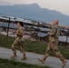 Two military personnel in camouflage uniforms and tan combat boots are jogging along a paved path at what appears to be a military installation or base. The facility is surrounded by security fencing with razor wire, and multiple low-profile buildings are visible in the background. A large mountain range is visible in the distance under a twilight or dawn sky.