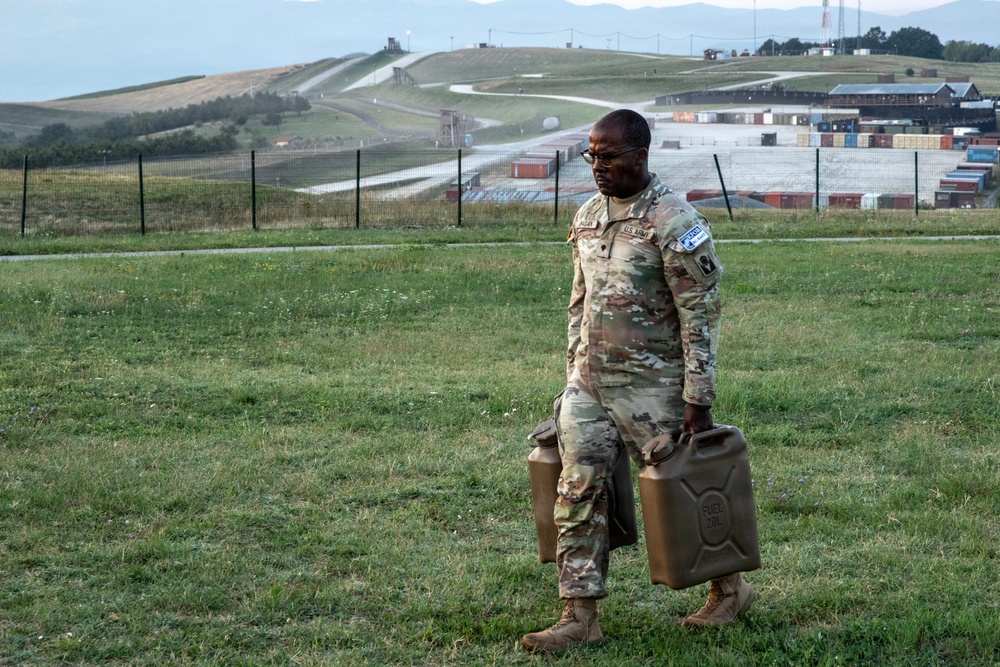 A U.S. Army soldier in OCP (Operational Camouflage Pattern) uniform is carrying two military fuel canisters across a grassy field at what appears to be a military installation or training base. The soldier is wearing glasses and has visible unit patches on the left sleeve. The background shows a large military compound with shipping containers, fencing, and a hillside with roads or runways.