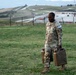 A U.S. Army soldier in OCP (Operational Camouflage Pattern) uniform is carrying two military fuel canisters across a grassy field at what appears to be a military installation or training base. The soldier is wearing glasses and has visible unit patches on the left sleeve. The background shows a large military compound with shipping containers, fencing, and a hillside with roads or runways.