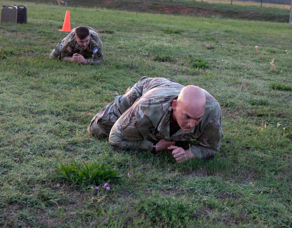 Two U.S. Army soldiers in OCP (Operational Camouflage Pattern) uniforms are performing a low crawl exercise on a grassy field. The foreground soldier is bald and has a name tape partially visible reading 'CKER', while a second soldier crawls in the background. This appears to be a military training exercise, likely a combat movement technique drill.