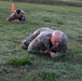 Two U.S. Army soldiers in OCP (Operational Camouflage Pattern) uniforms are performing a low crawl exercise on a grassy field. The foreground soldier is bald and has a name tape partially visible reading 'CKER', while a second soldier crawls in the background. This appears to be a military training exercise, likely a combat movement technique drill.