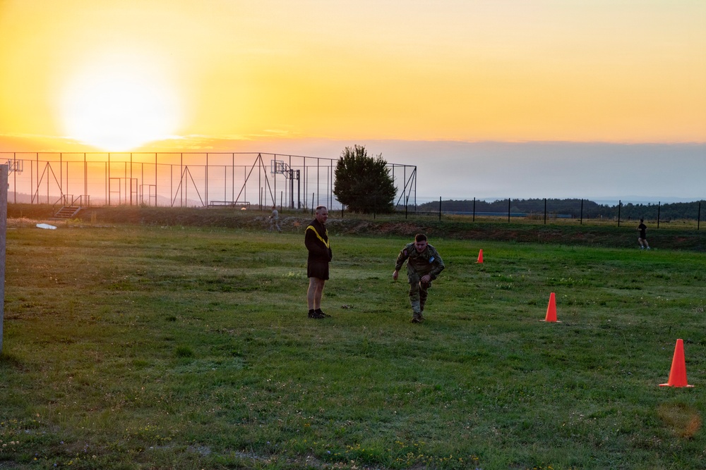 The image shows two individuals on a grassy field at sunrise or sunset, with one person in military camouflage uniform performing what appears to be a physical fitness drill near orange traffic cones. A second person wearing a dark hoodie and yellow sash or lanyard stands nearby, likely acting as an observer or evaluator. The scene appears to take place on a military training ground, with obstacle course structures and fencing visible in the background.