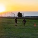 The image shows two individuals on a grassy field at sunrise or sunset, with one person in military camouflage uniform performing what appears to be a physical fitness drill near orange traffic cones. A second person wearing a dark hoodie and yellow sash or lanyard stands nearby, likely acting as an observer or evaluator. The scene appears to take place on a military training ground, with obstacle course structures and fencing visible in the background.