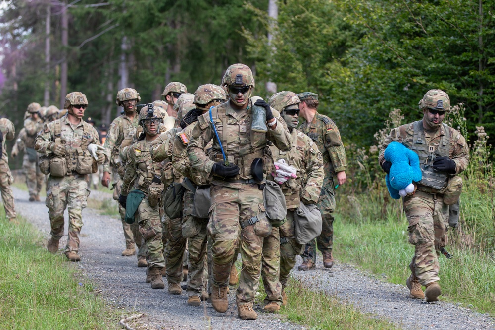 A group of soldiers in U.S. Army MultiCam/OCP camouflage uniforms are marching in a column along a gravel path through a forested area. The soldiers are fully equipped with tactical gear, helmets, and body armor. Notably, one soldier on the right is carrying a blue stuffed animal toy, and the lead soldier appears to be carrying miscellaneous civilian items, suggesting a training exercise involving simulated civilian casualty or humanitarian scenarios.