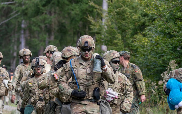 A group of soldiers in U.S. Army MultiCam/OCP camouflage uniforms are marching in a column along a gravel path through a forested area. The soldiers are fully equipped with tactical gear, helmets, and body armor. Notably, one soldier on the right is carrying a blue stuffed animal toy, and the lead soldier appears to be carrying miscellaneous civilian items, suggesting a training exercise involving simulated civilian casualty or humanitarian scenarios.