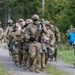 A group of soldiers in U.S. Army MultiCam/OCP camouflage uniforms are marching in a column along a gravel path through a forested area. The soldiers are fully equipped with tactical gear, helmets, and body armor. Notably, one soldier on the right is carrying a blue stuffed animal toy, and the lead soldier appears to be carrying miscellaneous civilian items, suggesting a training exercise involving simulated civilian casualty or humanitarian scenarios.