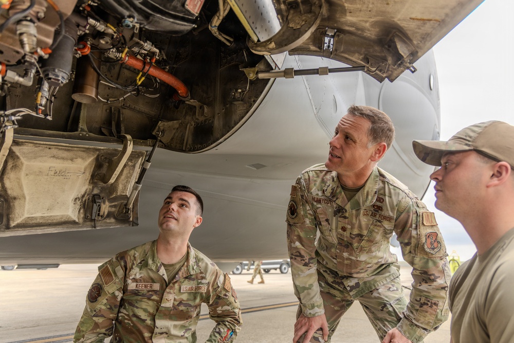 Three U.S. Air Force personnel are inspecting the underside of a military aircraft on a flight line. Two individuals are in full OCP (Operational Camouflage Pattern) uniforms with name tapes visible, while a third wears a tan cap. They appear to be examining engine or mechanical components beneath the aircraft fuselage.