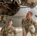 Three U.S. Air Force personnel are inspecting the underside of a military aircraft on a flight line. Two individuals are in full OCP (Operational Camouflage Pattern) uniforms with name tapes visible, while a third wears a tan cap. They appear to be examining engine or mechanical components beneath the aircraft fuselage.
