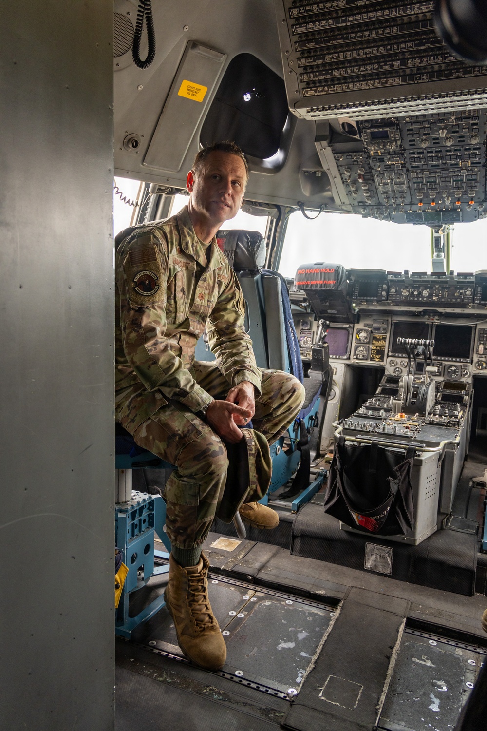 A U.S. Air Force service member in OCP (Operational Camouflage Pattern) uniform is seated in the cockpit doorway of a large military aircraft, likely a C-17 Globemaster III or similar transport aircraft. The individual is looking upward and to the side, with the aircraft's flight deck instrument panels and controls visible in the background. A 'NO HAND HOLD' warning label is visible on the pilot's seat headrest.
