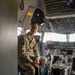 A U.S. Air Force service member in OCP (Operational Camouflage Pattern) uniform is seated in the cockpit doorway of a large military aircraft, likely a C-17 Globemaster III or similar transport aircraft. The individual is looking upward and to the side, with the aircraft's flight deck instrument panels and controls visible in the background. A 'NO HAND HOLD' warning label is visible on the pilot's seat headrest.