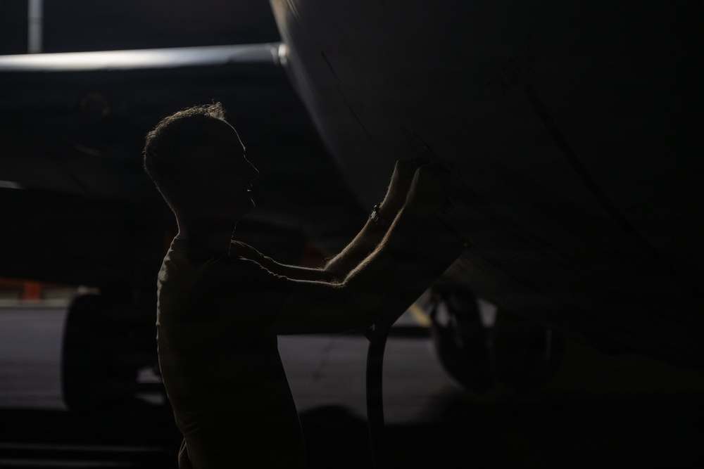 A silhouetted individual is performing maintenance or inspection work on the underside of a large aircraft in what appears to be a hangar or tarmac setting. The image is dramatically lit with strong contrast between the dark background and the partially illuminated figure and aircraft fuselage. The scene suggests a pre-flight check or routine mechanical inspection.