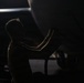 A silhouetted individual is performing maintenance or inspection work on the underside of a large aircraft in what appears to be a hangar or tarmac setting. The image is dramatically lit with strong contrast between the dark background and the partially illuminated figure and aircraft fuselage. The scene suggests a pre-flight check or routine mechanical inspection.