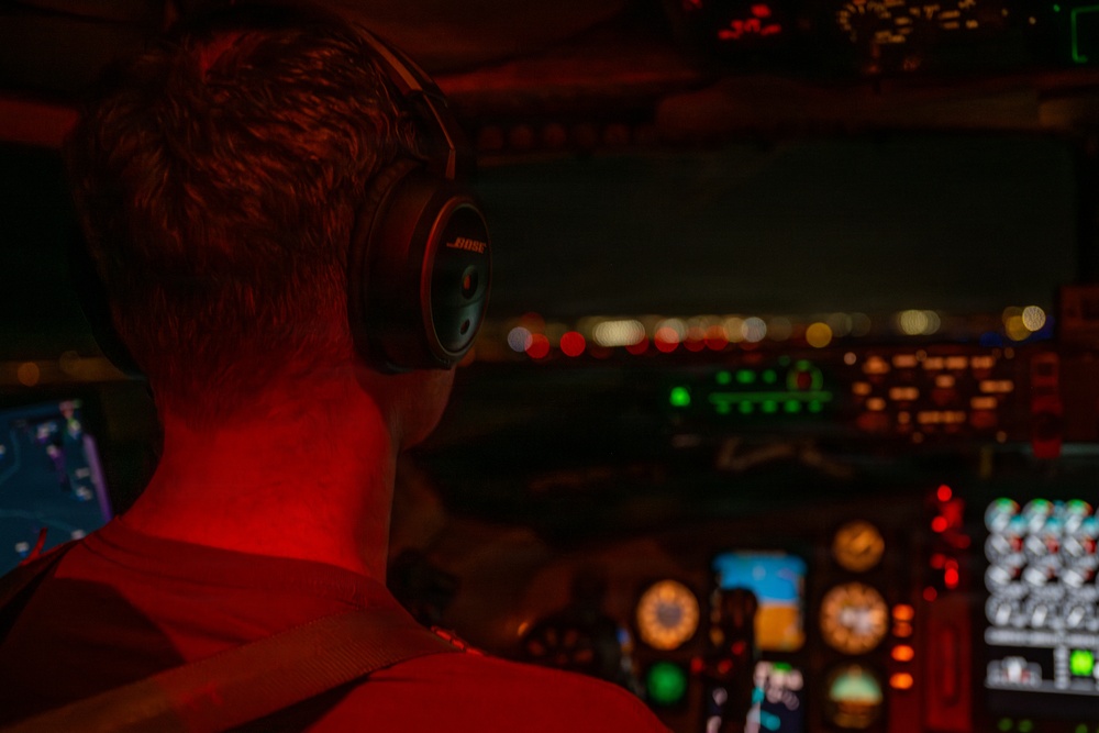 A pilot is seated in an aircraft cockpit during nighttime operations, viewed from behind. The cockpit is illuminated by red ambient lighting along with multicolored instrument panel displays. City lights are visible through the windscreen, suggesting the aircraft is on approach or taxiing at an airport.