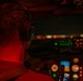 A pilot is seated in an aircraft cockpit during nighttime operations, viewed from behind. The cockpit is illuminated by red ambient lighting along with multicolored instrument panel displays. City lights are visible through the windscreen, suggesting the aircraft is on approach or taxiing at an airport.