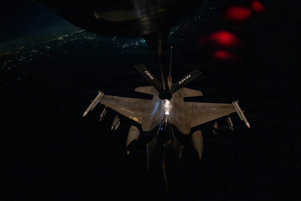 A night aerial refueling operation is captured showing a fighter jet (likely an F-16) receiving fuel from a tanker aircraft via a boom refueling system. The image is taken from the perspective of the tanker aircraft looking down at the receiver jet, with city lights visible in the background far below. The tanker's tail section displays the markings '196' and 'ARW' (Air Refueling Wing).