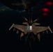 A night aerial refueling operation is captured showing a fighter jet (likely an F-16) receiving fuel from a tanker aircraft via a boom refueling system. The image is taken from the perspective of the tanker aircraft looking down at the receiver jet, with city lights visible in the background far below. The tanker's tail section displays the markings '196' and 'ARW' (Air Refueling Wing).