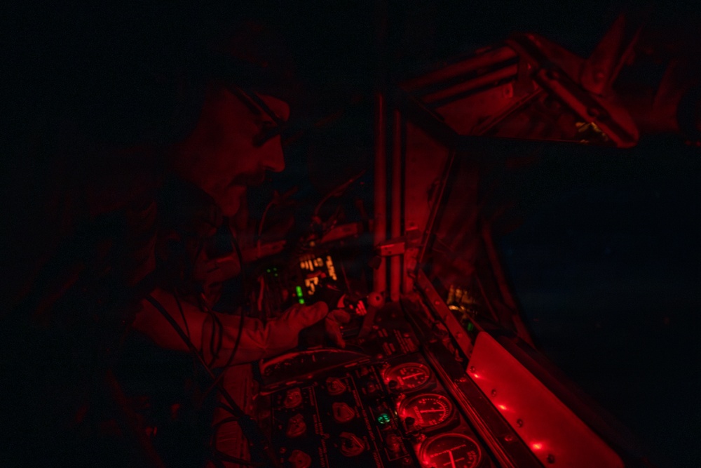 A military aircrew member is visible in a cockpit during nighttime operations, illuminated by red cockpit lighting. The red lighting is consistent with night vision preservation protocols used in military aviation. Multiple analog and digital instruments are visible on the instrument panel.