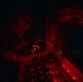 A military aircrew member is visible in a cockpit during nighttime operations, illuminated by red cockpit lighting. The red lighting is consistent with night vision preservation protocols used in military aviation. Multiple analog and digital instruments are visible on the instrument panel.