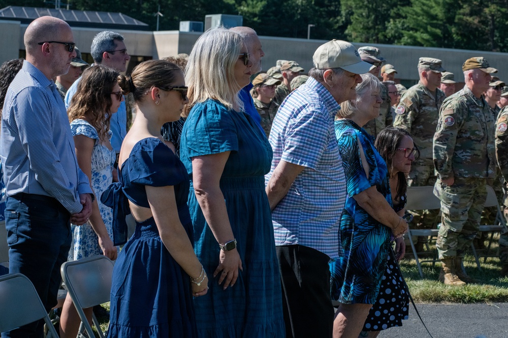 A military ceremony is taking place outdoors, with civilians standing in the foreground, likely family members of service members. In the background, a large formation of soldiers in camouflage uniforms (OCP pattern) is assembled. The civilians appear to be observing or participating in a formal military event such as a deployment ceremony, homecoming, or change of command.