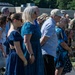 A military ceremony is taking place outdoors, with civilians standing in the foreground, likely family members of service members. In the background, a large formation of soldiers in camouflage uniforms (OCP pattern) is assembled. The civilians appear to be observing or participating in a formal military event such as a deployment ceremony, homecoming, or change of command.