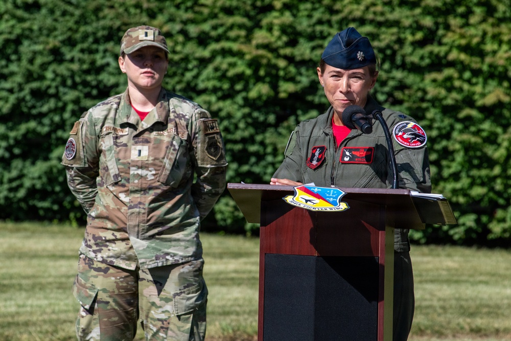 Two female U.S. Air Force personnel are participating in an outdoor military ceremony. The officer at the podium, wearing a flight suit and blue service cap with a lieutenant colonel oak leaf insignia, is delivering remarks at what appears to be an official event. A second service member in OCP (Operational Camouflage Pattern) uniform stands at attention in the background.