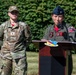 Two female U.S. Air Force personnel are participating in an outdoor military ceremony. The officer at the podium, wearing a flight suit and blue service cap with a lieutenant colonel oak leaf insignia, is delivering remarks at what appears to be an official event. A second service member in OCP (Operational Camouflage Pattern) uniform stands at attention in the background.