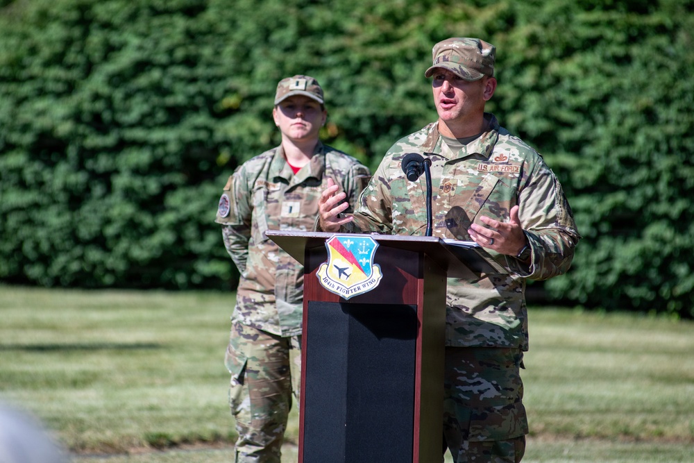 Two U.S. Air Force personnel in OCP (Operational Camouflage Pattern) uniforms are participating in an outdoor military ceremony or briefing. The individual at the podium is speaking into a microphone, while a second service member stands at attention in the background. The podium displays the insignia of the 104th Fighter Wing.