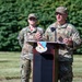 Two U.S. Air Force personnel in OCP (Operational Camouflage Pattern) uniforms are participating in an outdoor military ceremony or briefing. The individual at the podium is speaking into a microphone, while a second service member stands at attention in the background. The podium displays the insignia of the 104th Fighter Wing.